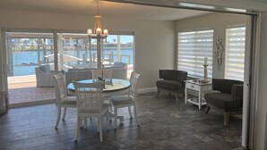 Un espace repas offrant une vue sur un port de plaisance, avec une table ronde entourée de chaises, un lustre et une grande fenêtre équipée de stores.