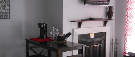 A cozy living room with a fireplace, a television mounted above it, and a small table with a coffee maker and decorative items.