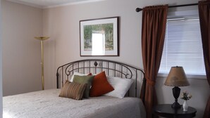 Un lit bien fait, avec une couette blanche et plusieurs oreillers, un tableau encadré au mur, une lampe sur une table de chevet et une fenêtre avec des rideaux.