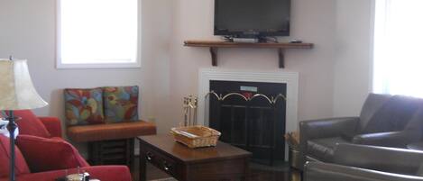 A living room with a fireplace, a television mounted above it, a red sofa, a grey armchair, and a coffee table.