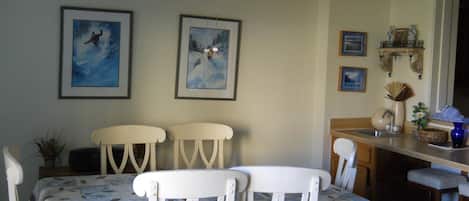 A dining area with a table and chairs, framed artwork on the wall, and a kitchen counter with a sink and decorative items.