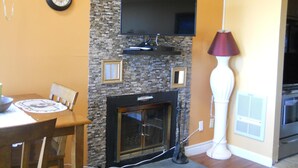 Ein Wohnzimmer mit Kamin, einem flachbildfernseher darüber, einem Esstisch mit Stühlen und einer Lampe.