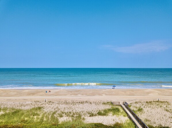 Una playa con un paseo marítimo de madera que lleva al agua, dunas arenosas y un cielo azul despejado.