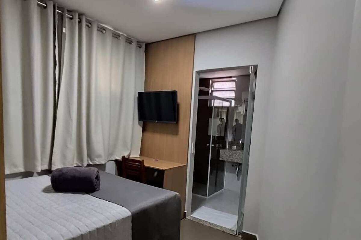 Habitación de hotel con cama, escritorio, televisión y baño con puerta de vidrio.