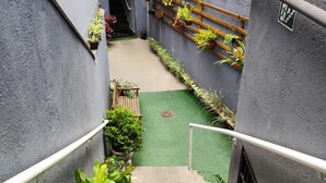 Eine enge Treppe führt zu einem kleinen Innenhof mit einem Rasenbereich aus künstlichem Grün, umgeben von grauen Mauern und verschiedenen Topfpflanzen.