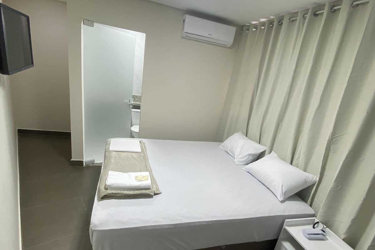 Una habitación de hotel con cama, mesita de noche con lámpara, televisor montado en la pared y equipo de aire acondicionado.