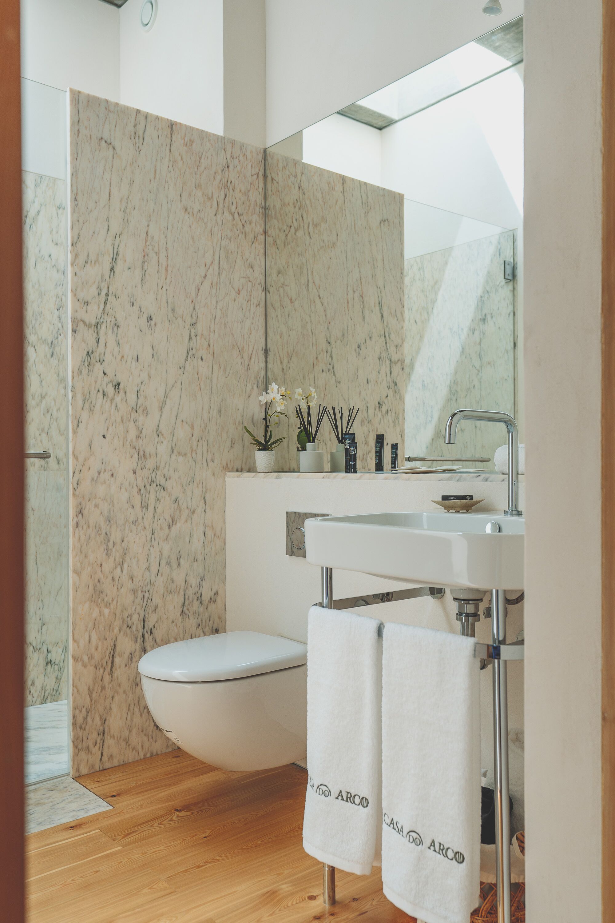 Une salle de bain moderne avec un mur en marbre, un lavabo blanc et des toilettes. Des serviettes sont suspendues sur un porte-serviettes et une petite plante est posée sur le lavabo.