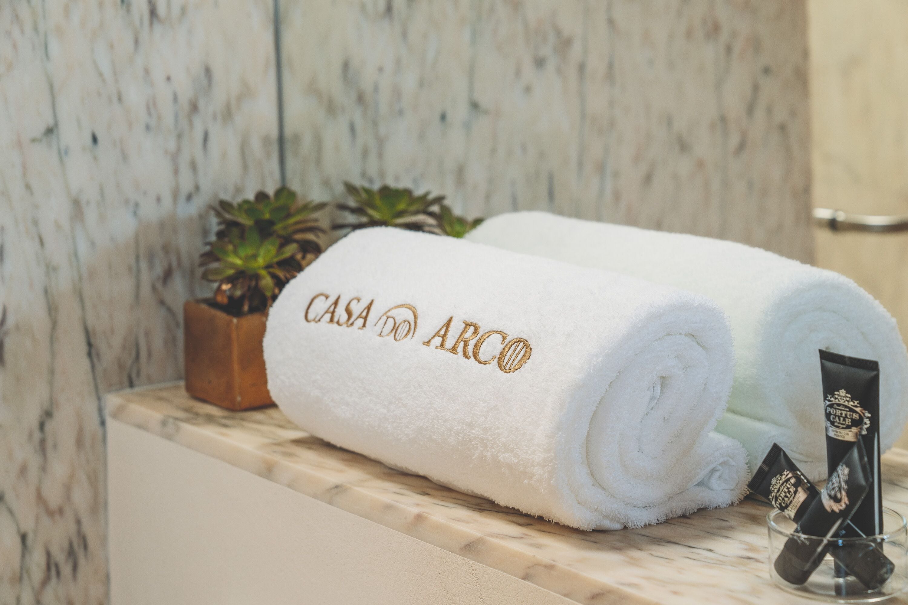 Deux serviettes blanches ornées du motif « CASA DO ARCO » brodé, une petite plante en pot et un récipient en verre contenant un tube noir.