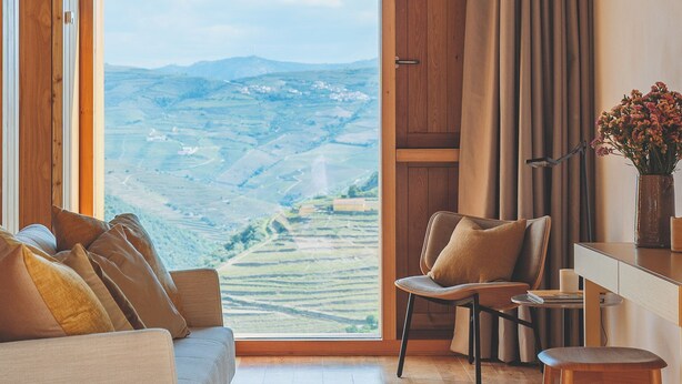 Una habitación moderna con suelo de madera, un sofá, una silla, un escritorio y vistas a un paisaje a través de un ventanal grande.