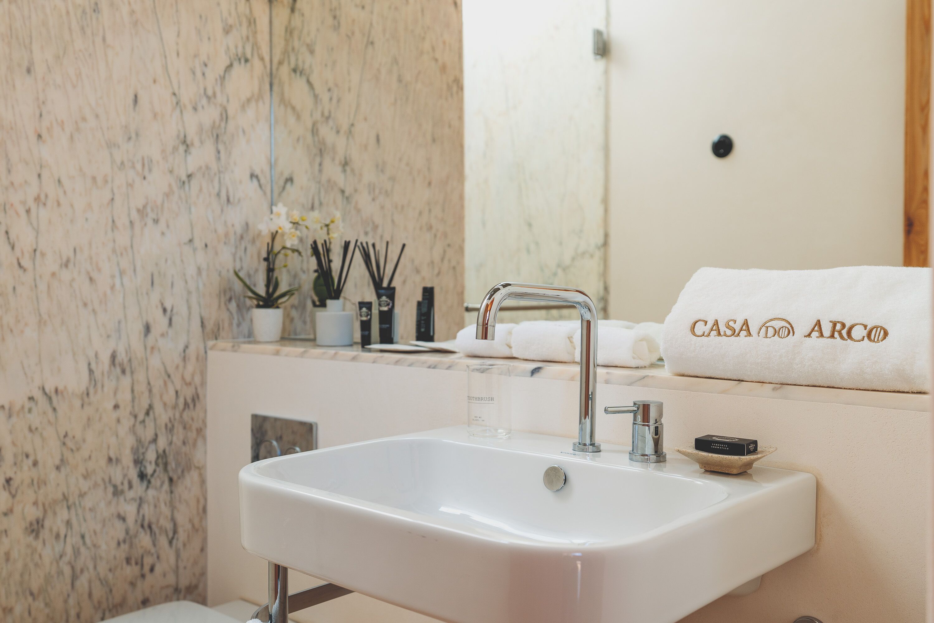 A bathroom with a white sink, a mirror, and a towel rack with a towel labeled "CASA DO ARCO".