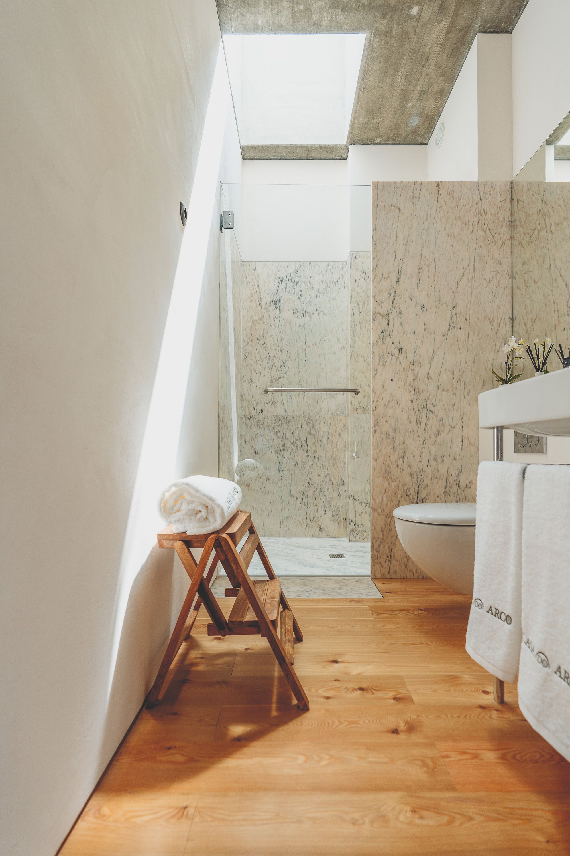 Un baño moderno con tragaluz, suelo de madera, pared de ducha de mármol y lavabo blanco.