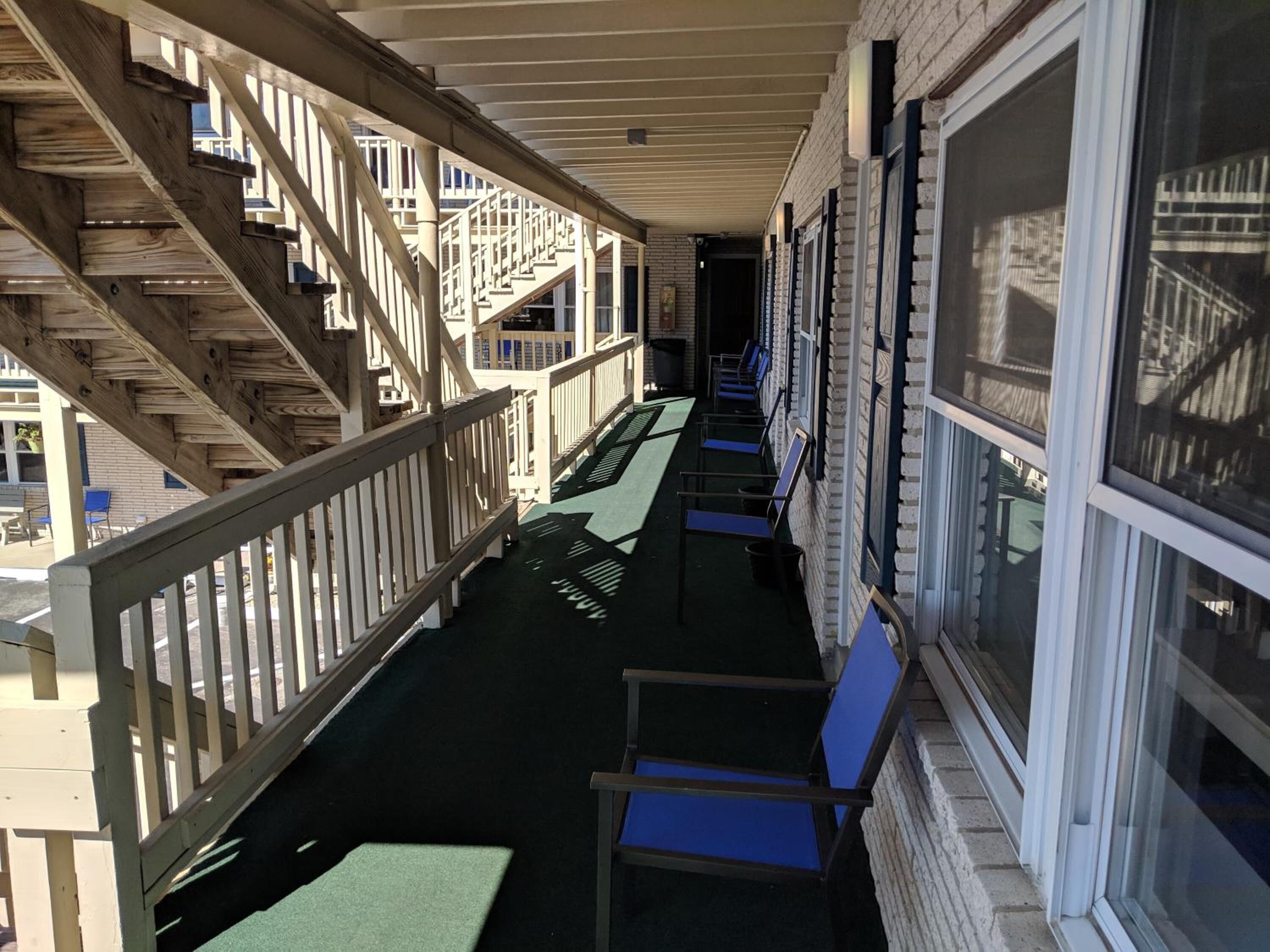 A covered outdoor area with a wooden staircase, rows of blue chairs, and a brick building with large windows.