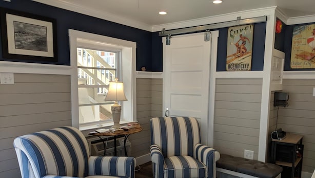 A room with striped armchairs, a bench, a lamp, a window with a view of a building, and a door with a poster. - Crystal Sands Motel (Ocean City)