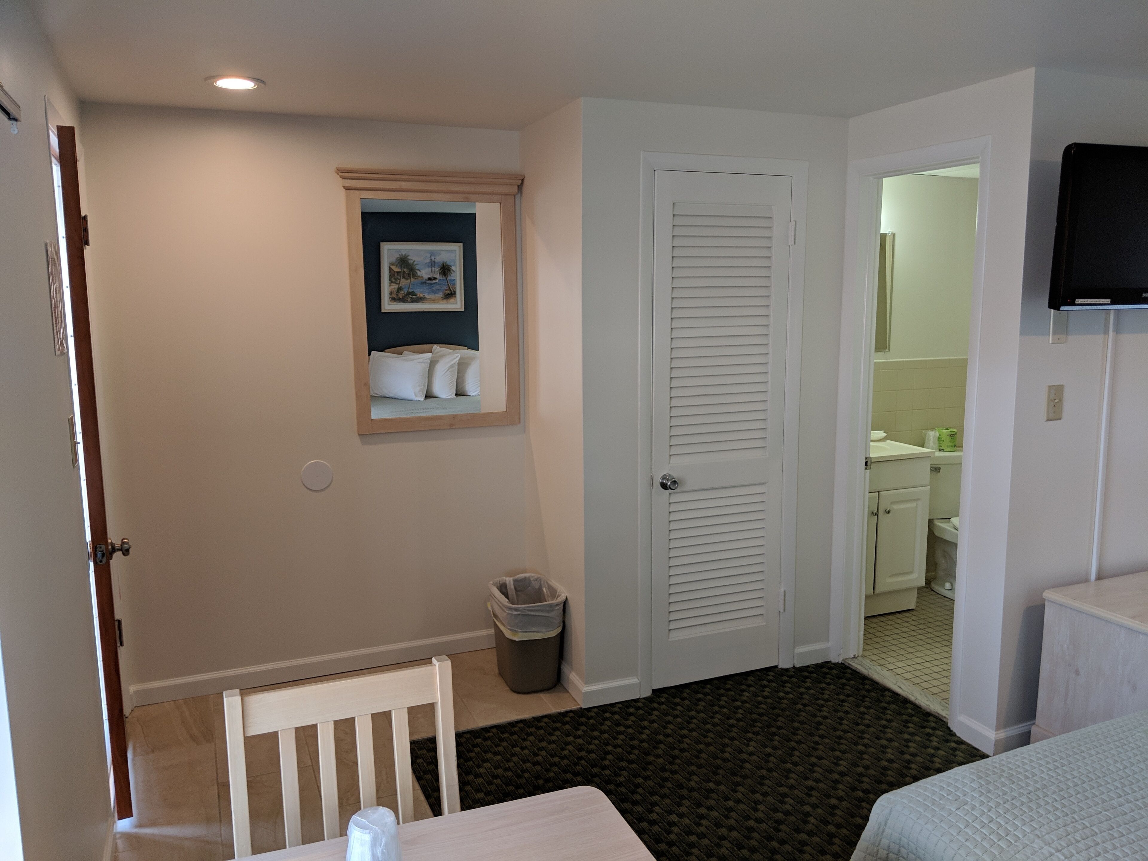 Habitación de hotel con cama, una mesita, una silla, un cubo de basura, un marco de foto y un baño visible a través de una puerta abierta.