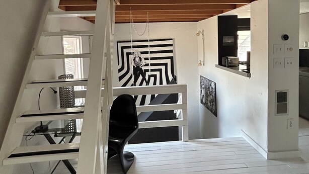 Ein geräumiger Loft mit hoher Decke, freiliegenden Holzbalken, einer Treppe mit weißen Geländern und einem schwarz-weißen abstrakten Gemälde an der Wand.