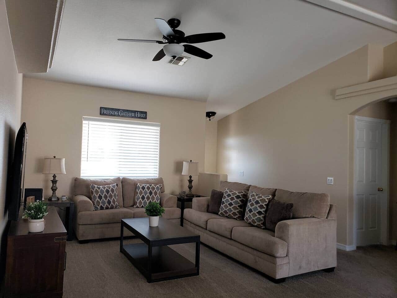 A living room with a two-seater sofa, a coffee table, a ceiling fan, and a sign that reads "Friends Gather Here".