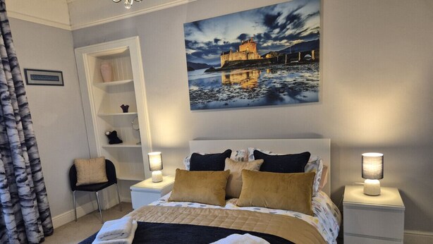 A bedroom with a large bed, two bedside tables with lamps, a chair, a shelf, and a painting of a castle.