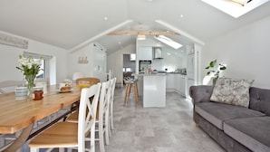 A modern kitchen with white cabinetry and a central island, a dining area with a wooden table and white chairs, and a grey sofa with a floral pillow. - Charming Cottage, Wi-Fi, Parking, Pets Accepted (Newquay)