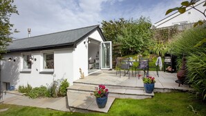 A white building with a grey roof, a glass door entrance, and a patio area with outdoor seating. - Charming Cottage, Wi-Fi, Parking, Pets Accepted (Newquay)