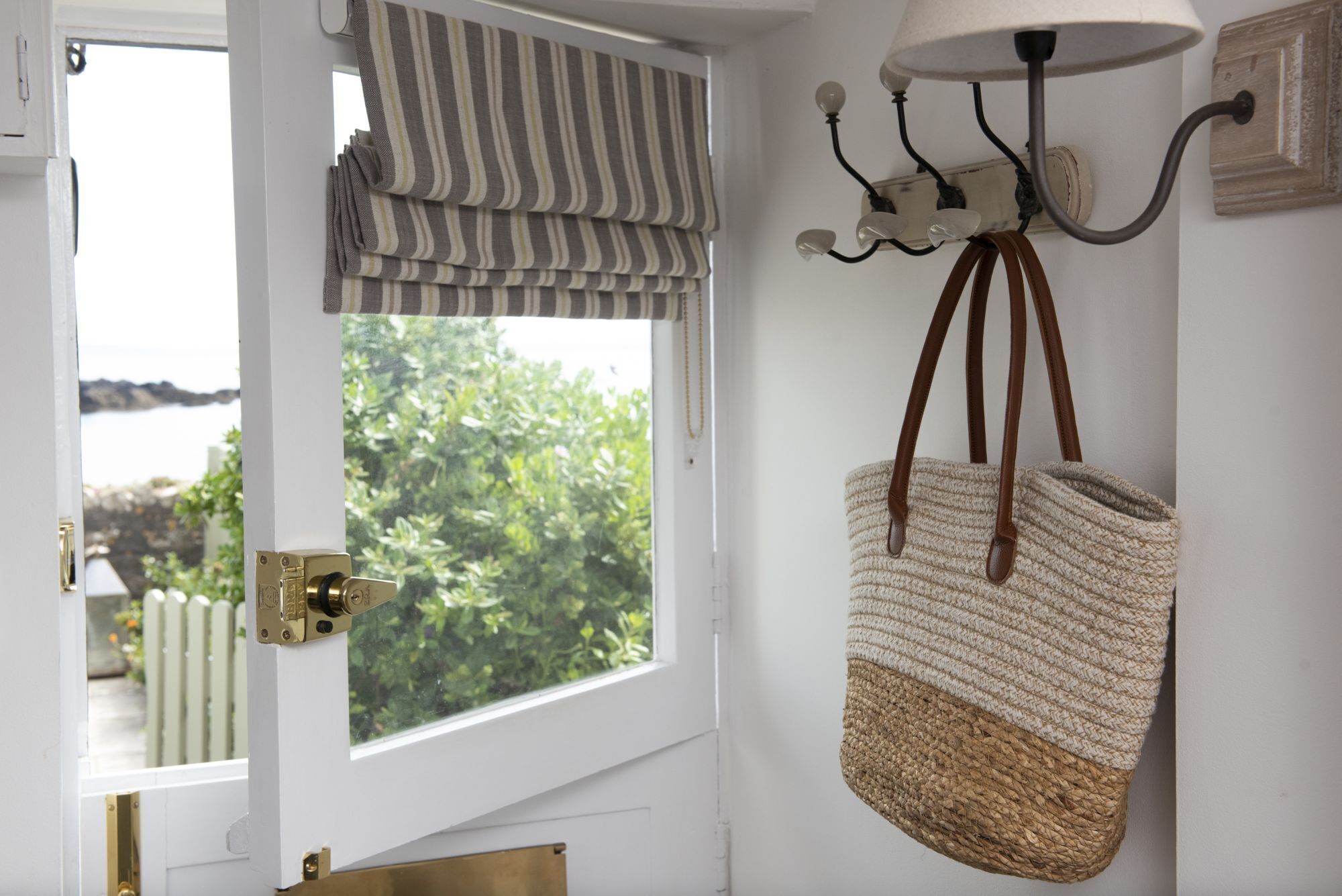 Une pièce avec une fenêtre, une porte dotée d’une poignée en laiton, une lampe fixée au mur et un sac en tissu suspendu au mur.