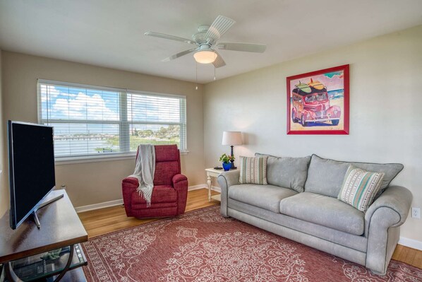 A living room with a red armchair, a grey sofa, a ceiling fan, a painting, and a view through the window.