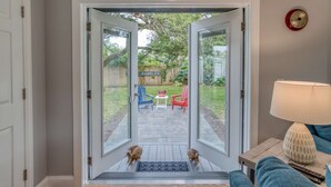 Blick durch eine Glastür in einen Hinterhof mit einer Holzterrasse, zwei Stühlen und einem kleinen Tisch.