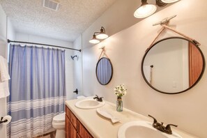 A bathroom with a shower curtain, two sinks, a mirror, and a vase with flowers. - Barefoot Hideaway (St. Augustine)