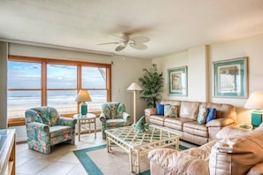 A living room with a view of the beach, a ceiling fan, and a sofa with floral cushions.