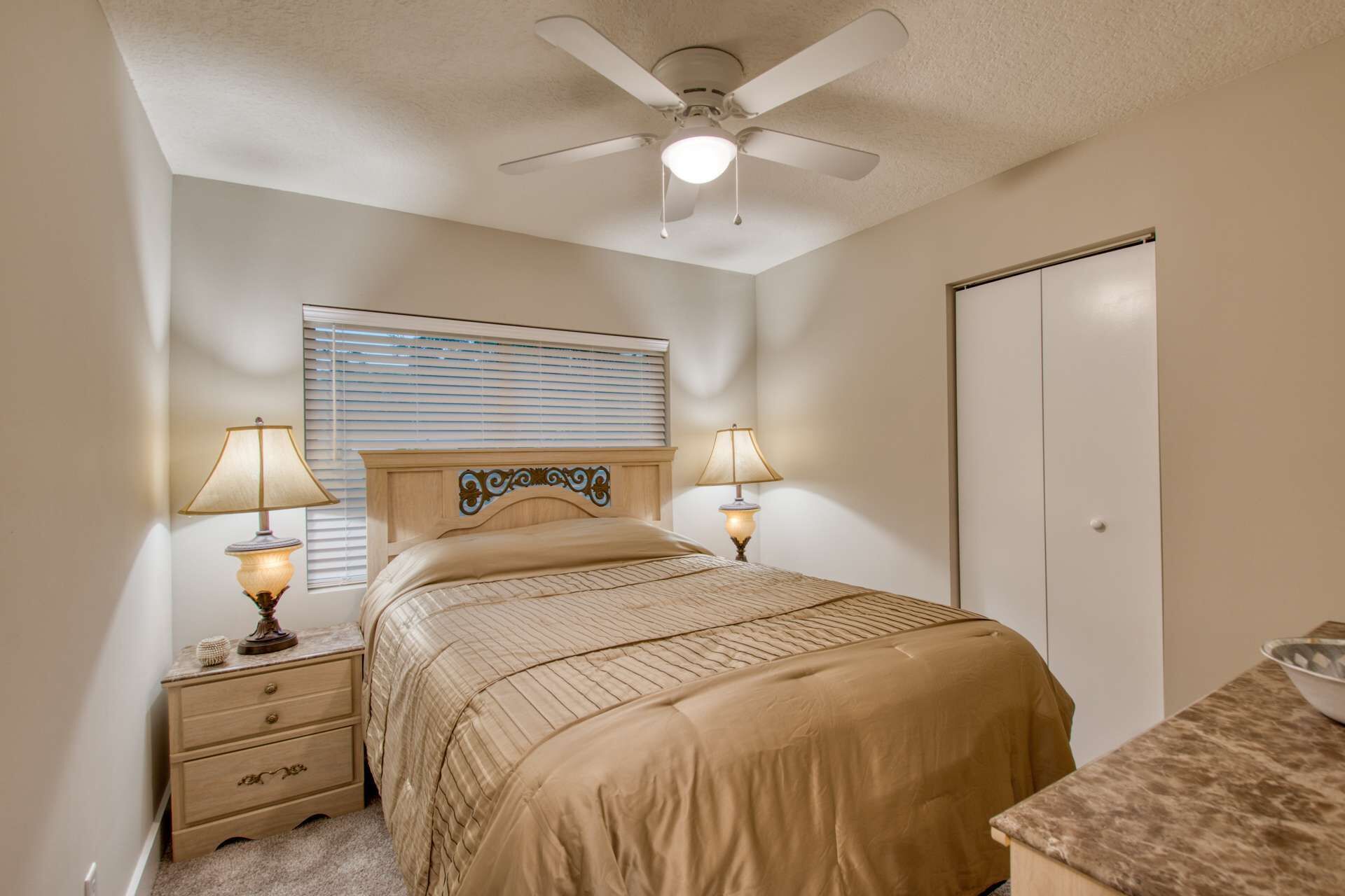 A bedroom with a bed, two bedside lamps, a ceiling fan, a window with blinds, and a wardrobe.