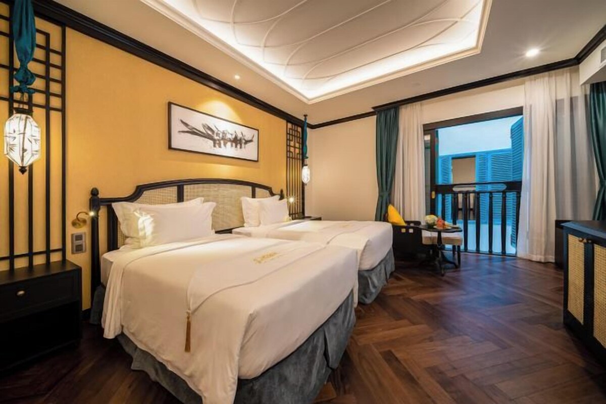 A hotel room with two beds, a balcony with curtains, a desk with a chair, and a wall-mounted clock.