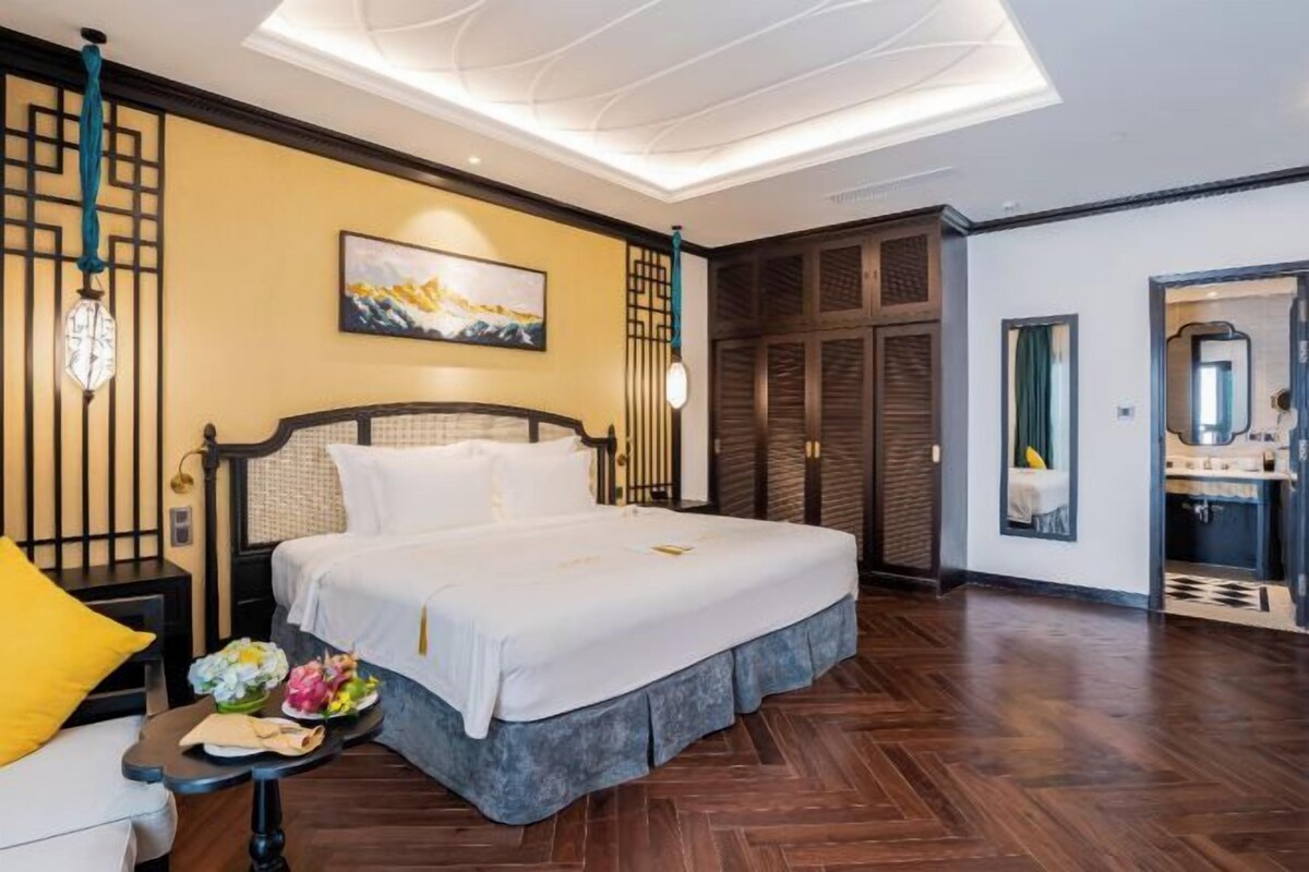 A hotel room with a large bed, a small table with a flower arrangement, a painting on the wall, and a traditional wooden door.