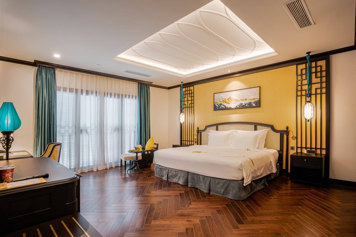 Une chambre d’hôtel avec un grand lit, un bureau, une chaise, une table avec une lampe et une fenêtre avec des rideaux.