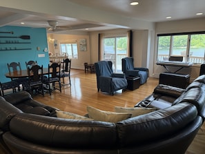 A spacious living room with a wooden floor, a black leather sofa, a blue armchair, a dining table with chairs, and a piano.