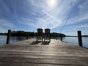 A wooden pier with two Adirondack chairs facing a calm lake, a fishing rod, and a clear sky.