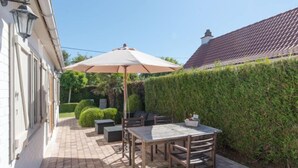 Eine Terrasse mit Tisch und Stühlen, einem großen Sonnenschirm und einem Hecken.
