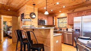 A wooden cabin interior with a kitchen featuring a central island, bar stools, a microwave, and a refrigerator. - Luxe Pool Cabin Near Dollywood & Top Attractions (Pigeon Forge)