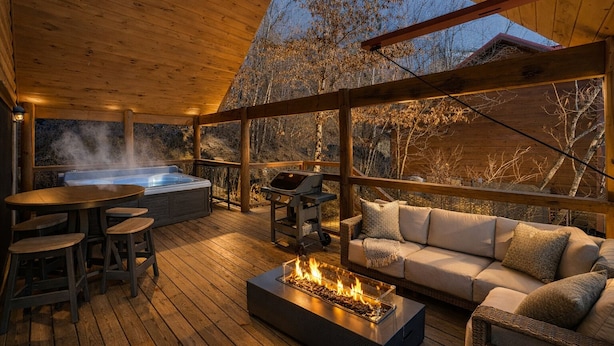A wooden deck with a hot tub, grill, and fire pit, surrounded by a wooden fence and overlooking a forested area. - Luxe Pool Cabin Near Dollywood & Top Attractions (Pigeon Forge)