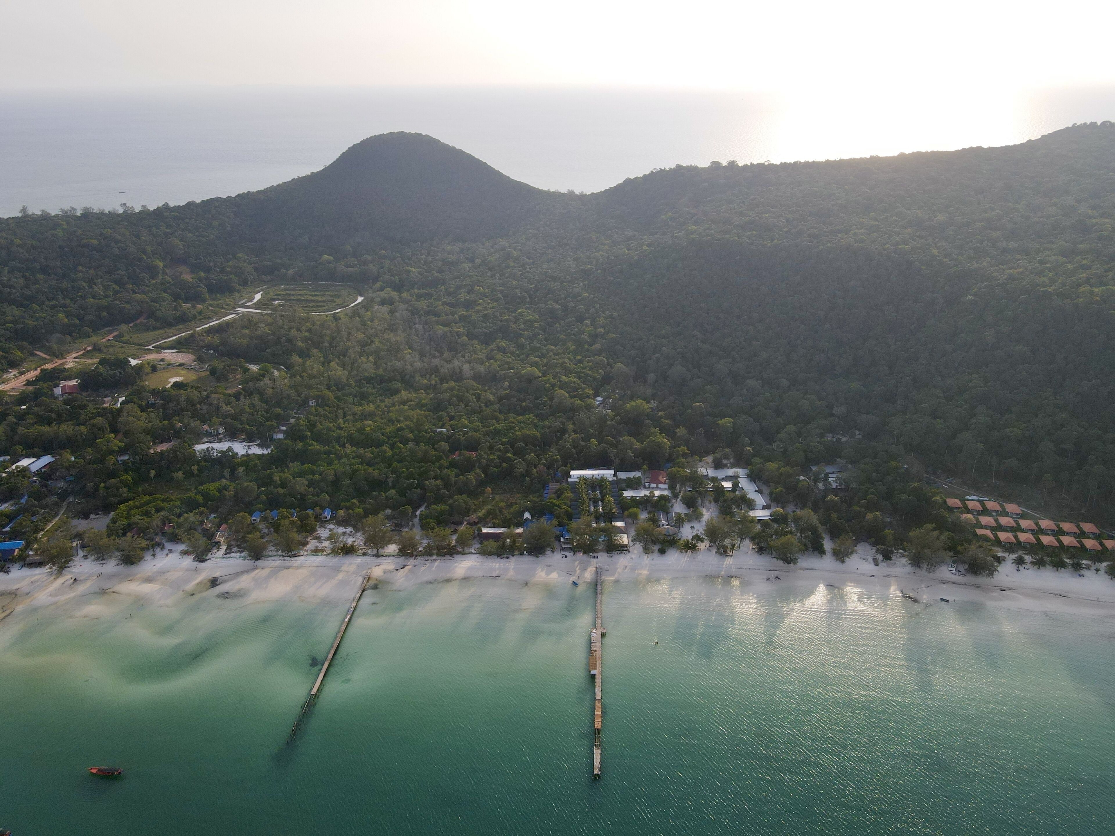 An aerial view of a coastal area with a beach, a pier, and a mountain in the background.