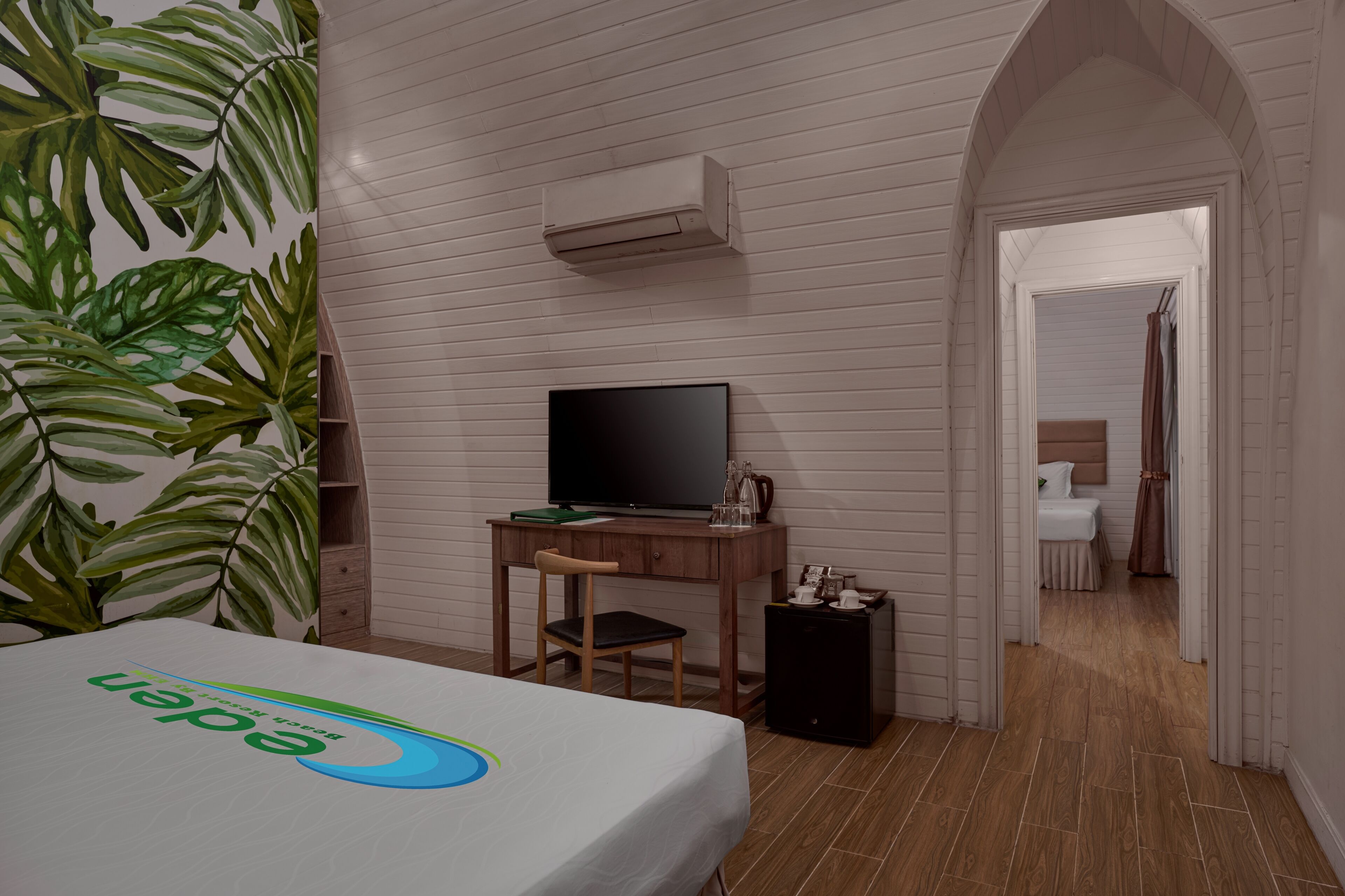 A hotel room with a bed, a desk with a TV, a small cabinet, and a plant.