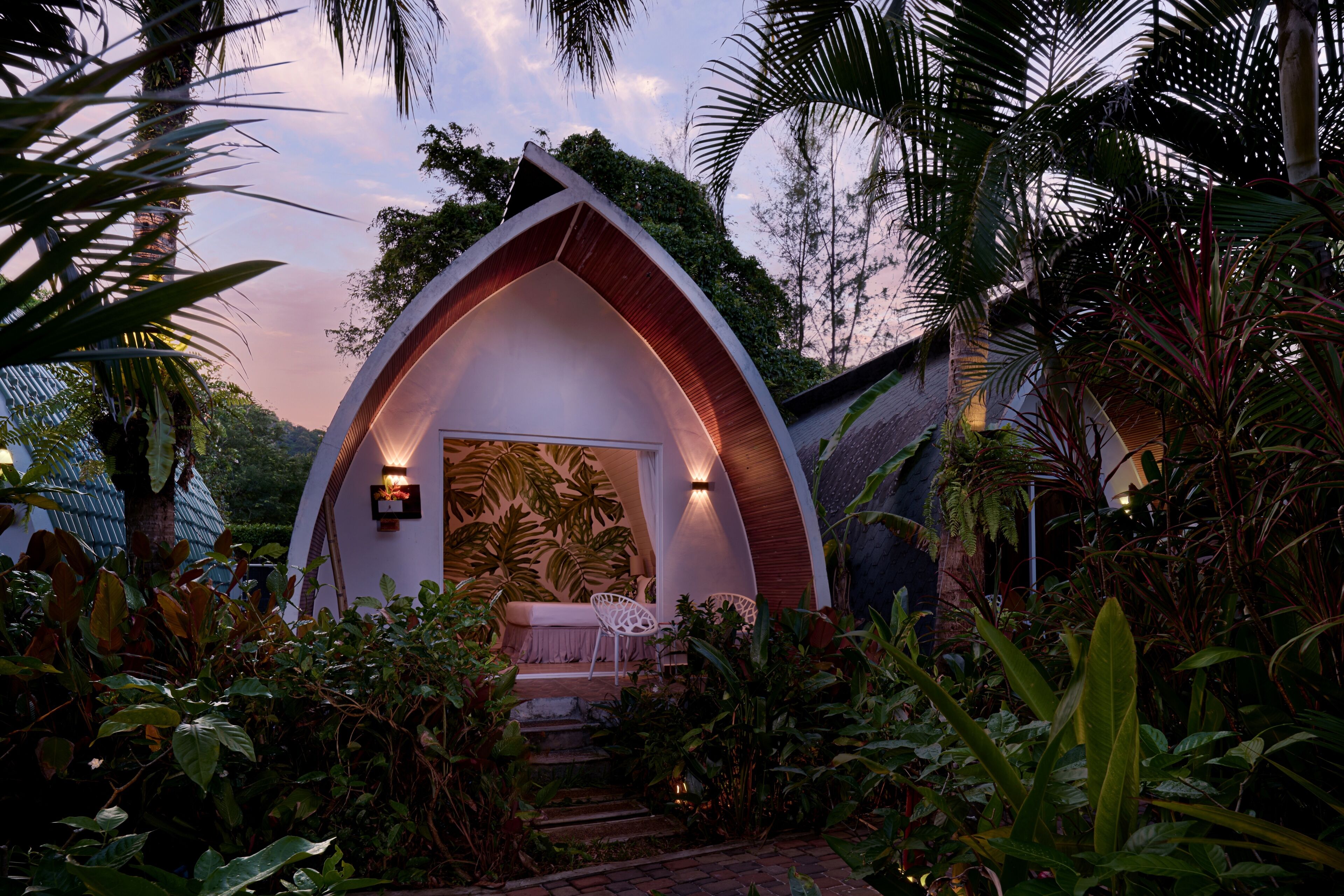 A small, white, arched structure with a wooden exterior and a bench inside, surrounded by lush greenery.