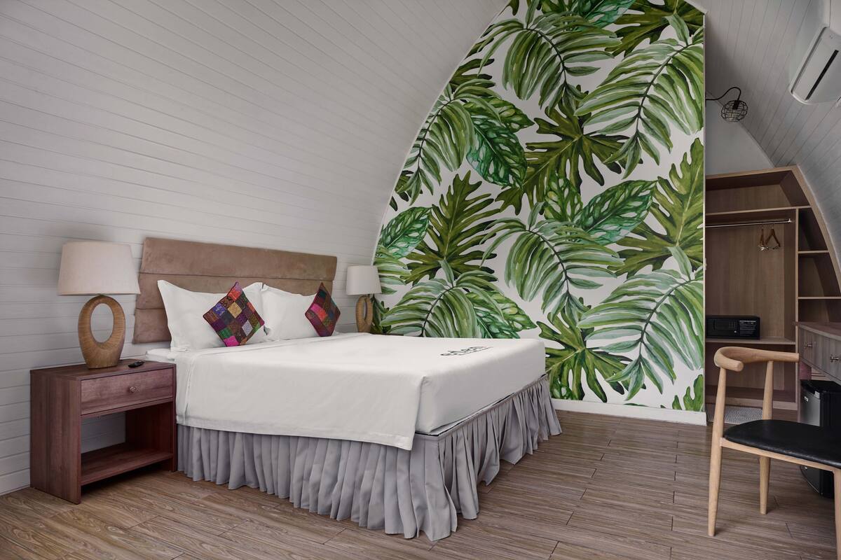 Un dormitorio con cama, mesita de noche, lámpara y una pared con estampado de hojas tropicales.