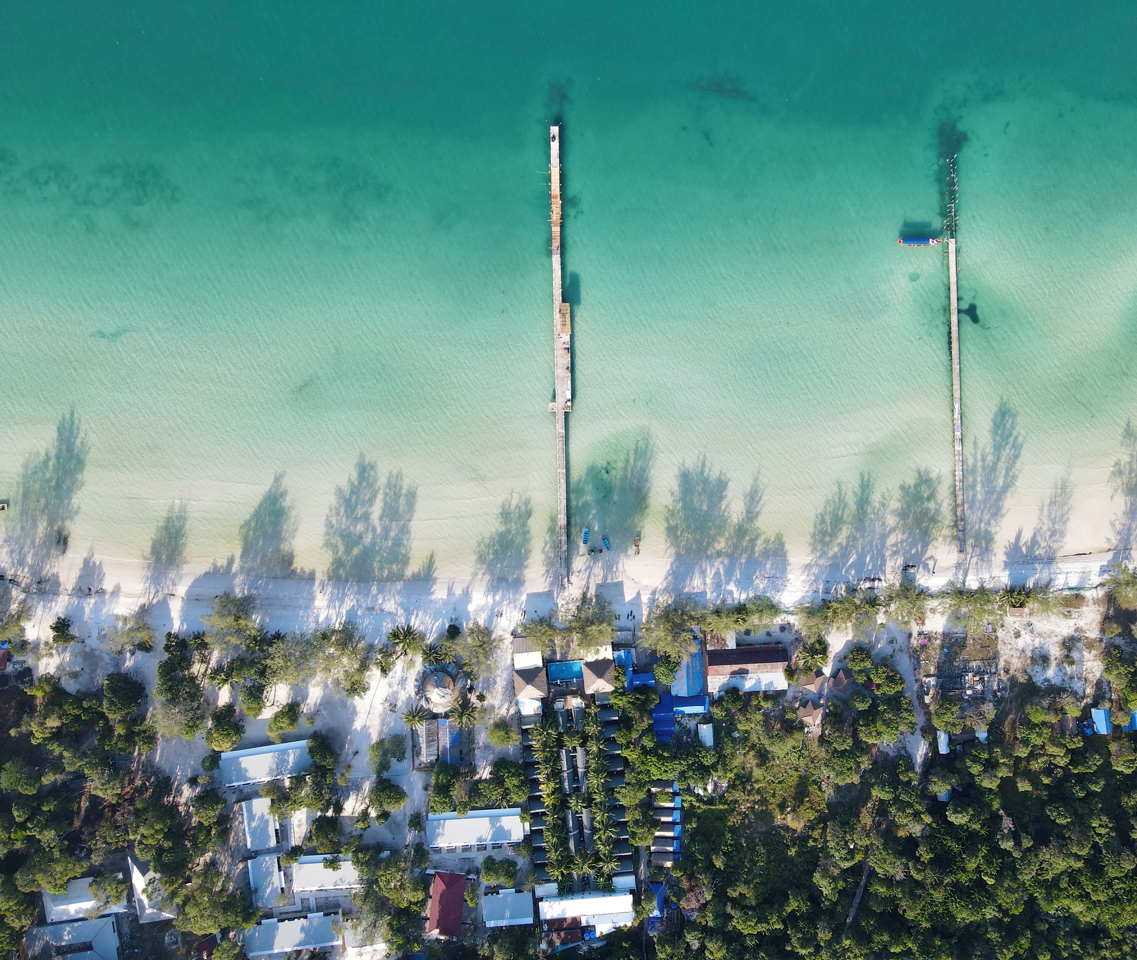 An aerial view of a coastal area with buildings, a pier extending into the water, and dense vegetation.