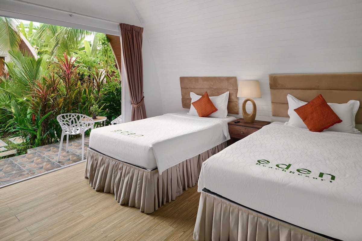 Una habitación con dos camas, una mesita y una silla. Las camas tienen una colcha blanca con el logo 'eden beach resort'. Hay una ventana con cortinas y vistas al exterior, lleno de vegetación.