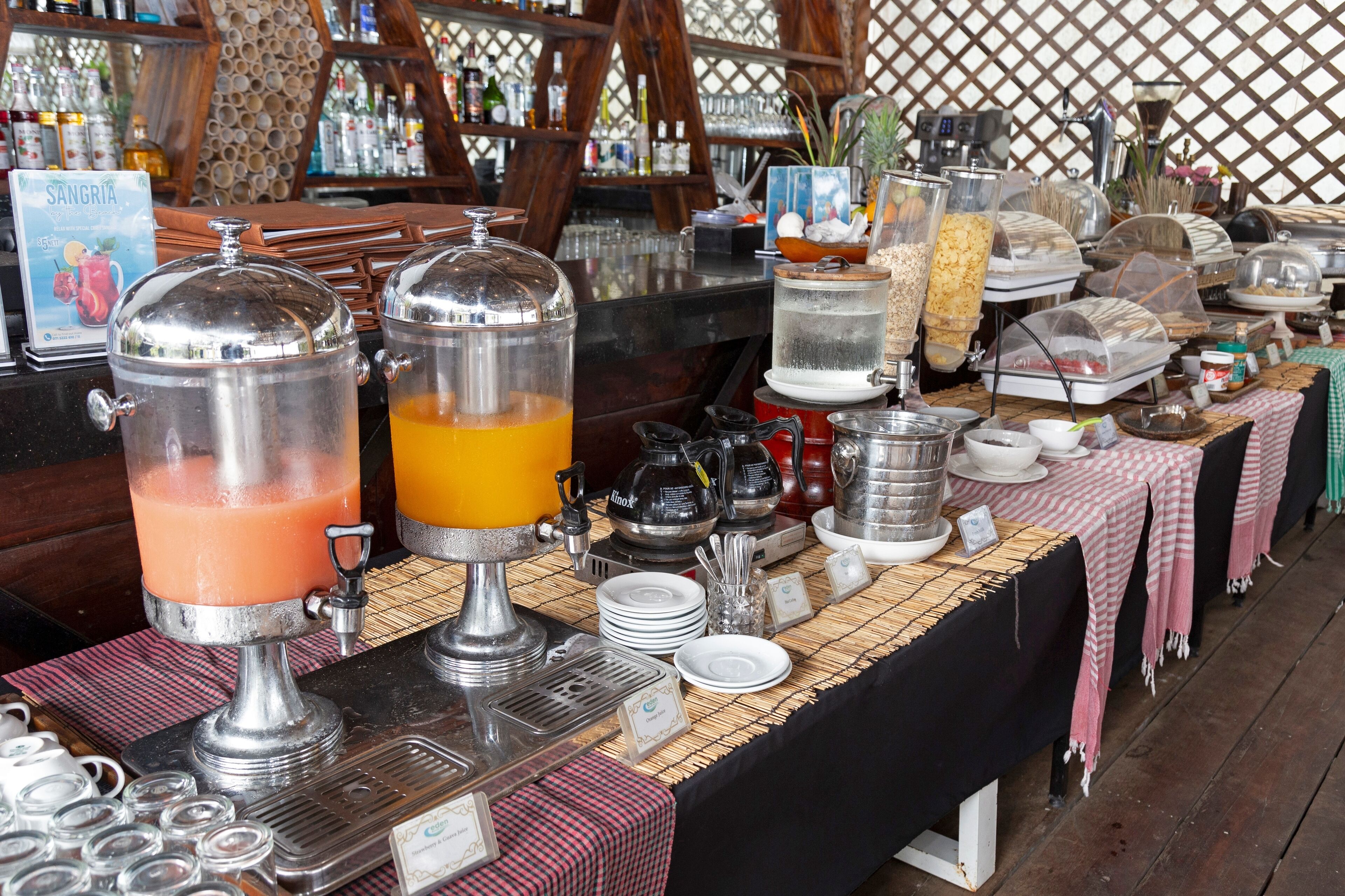 A buffet setup with various food and drink dispensers, including a large orange juice dispenser and a pink smoothie dispenser.