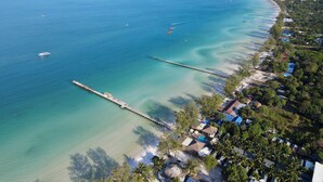 An aerial view of a coastal area with a pier extending into the sea, surrounded by residential buildings and a small island in the background. - Eden Beach Resort by EHM (Koh Rong Sanloem)