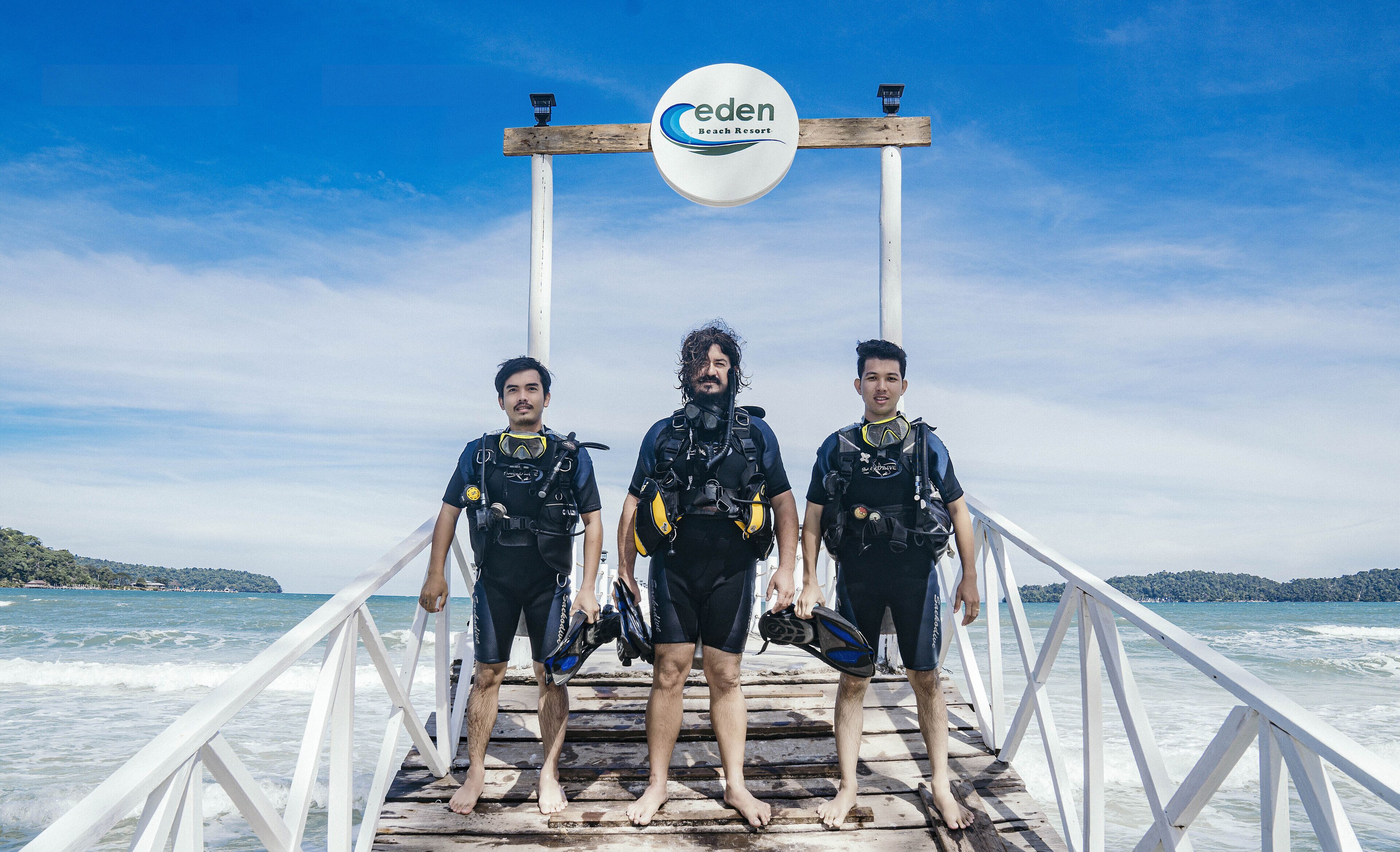 Drie duikers staan op een houten pier, met een bord erboven met daarop 'eden Beach Resort'.