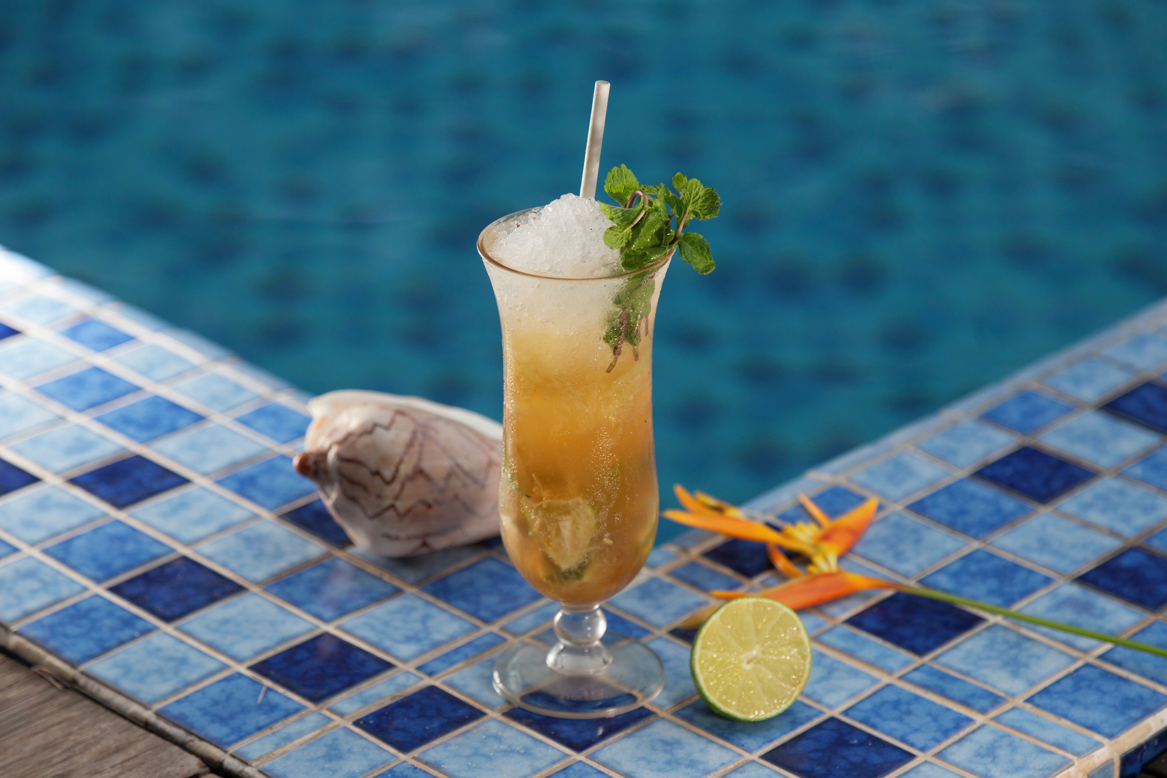 A cocktail glass with ice, garnished with a mint leaf, next to a lime wedge on a tiled surface by a pool.