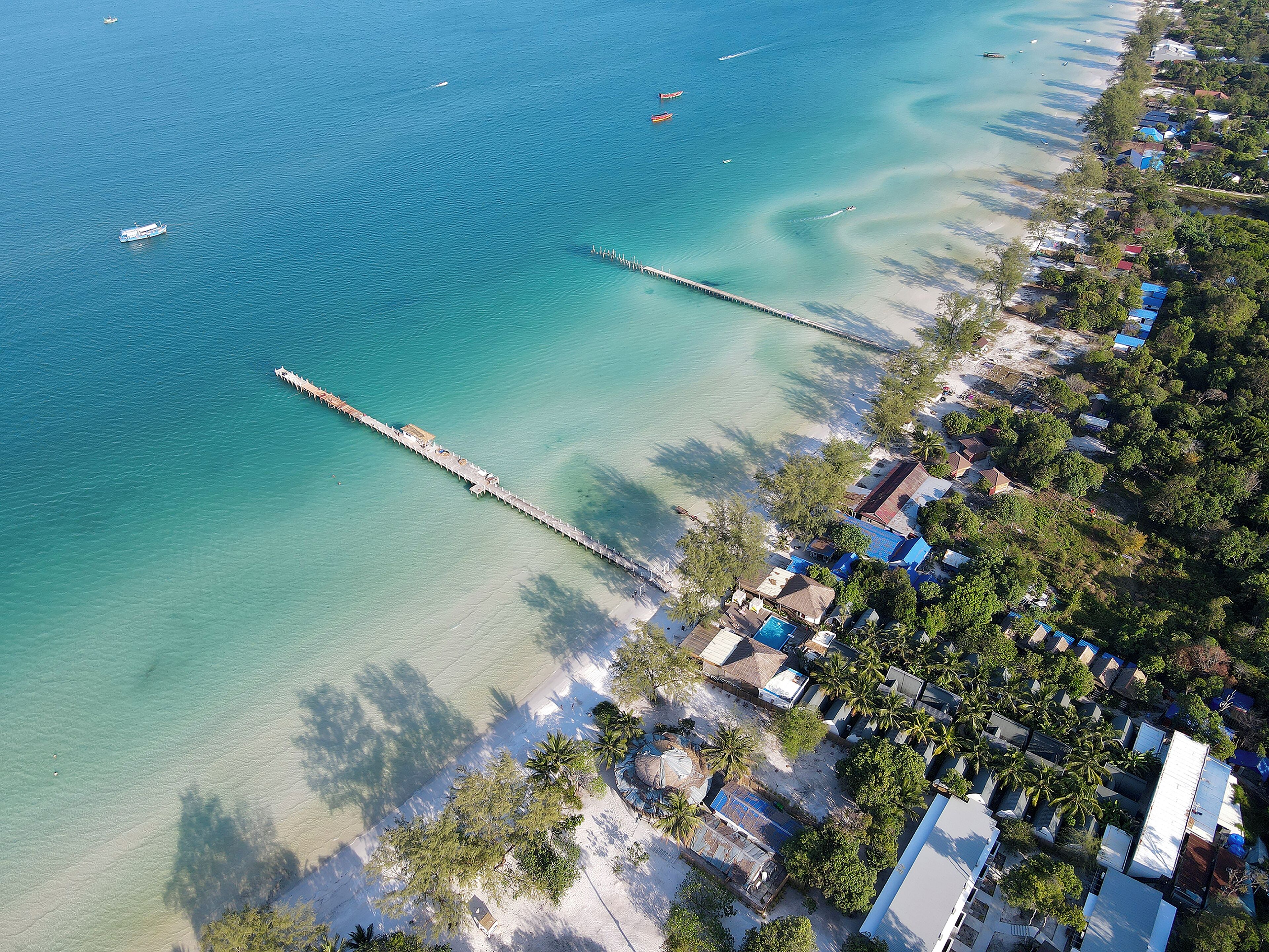 Vista aérea de una zona costera con un muelle que se adentra en el agua, rodeada de edificios residenciales y palmeras.