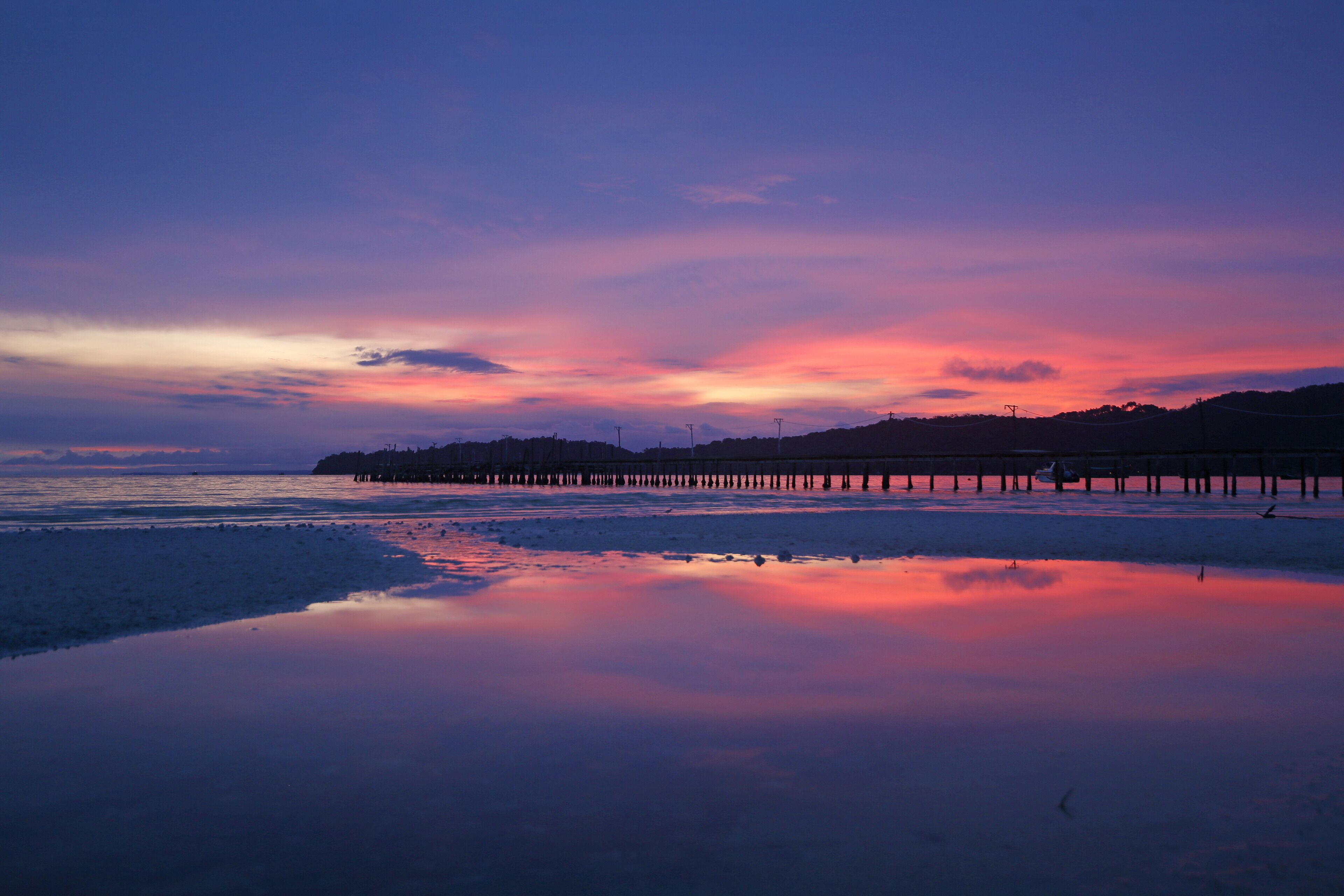 A beach with a pier extending into the water, reflecting the sunset colors.