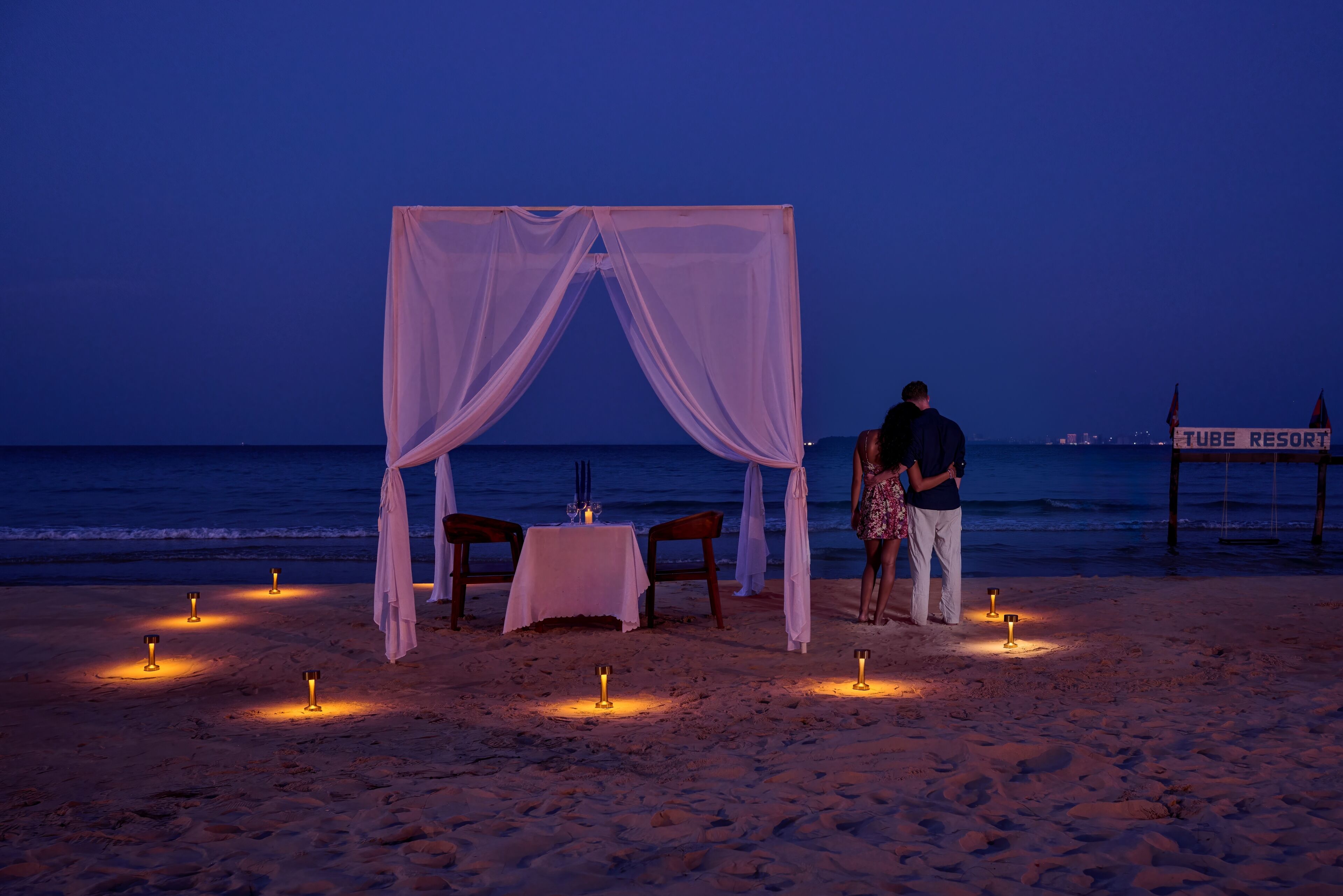A beachside tent with a table and chairs, candles, and a sign for 'Tube Resort.'